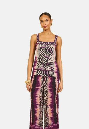 Femme portant un haut sans manches et un pantalon large avec un imprimé zèbre abstrait violet, noir et beige, debout devant un fond uni clair.