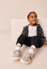 Hall halli teksari jakk, valge t-särk, mustad püksid ja hallid tossud Velcro rihmaga. Valged sukad pruunide triipudega tekstuuriga. Istuv poos.