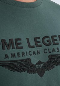 Grøn bomulds-t-shirt med sort broderet tekst "PME LEGEND AMERICAN CLASSIC" og vinge-grafik på forsiden. Korte ærmer, rund hals.