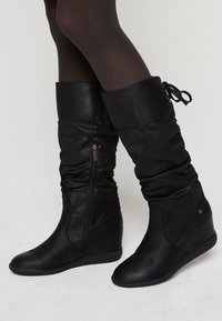 Stivali a zeppa neri a metà polpaccio con cerniera laterale, fusto arricciato, cima piegata e fiocco sul retro, indossati con calze nere trasparenti su gambe incrociate.
