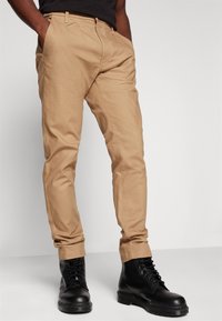 Chinos beige con un corte slim, ligero brillo y bolsillos laterales, combinados con botas negras de cordones con suela resistente y punta redonda.
