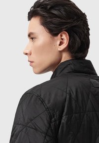 Czarna pikowana kurtka z wysokim kołnierzem, ozdobiona geometrycznym wzorem i metką z tyłu, podkreślająca elegancką, gładką teksturę.