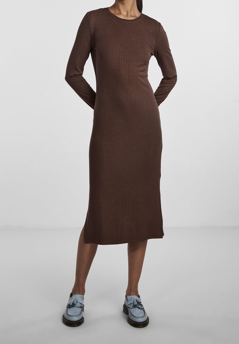 Femme portant une robe midi marron à manches longues côtelée, associée à des mocassins bleu clair avec des pampilles, debout devant un fond uni.