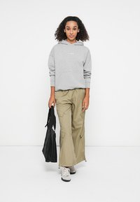 Grauer Kapuzenpullover mit einer vorderen Tasche, kombiniert mit beigen Cargo-Hosen mit Seitentaschen. Eine schwarze Tragetasche und graue Sneaker runden den Look ab.