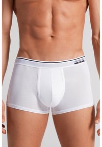 Mannelijk bovenlichaam met witte, aansluitende boxershorts met een merkband en een voorzak, staand tegen een neutrale achtergrond.