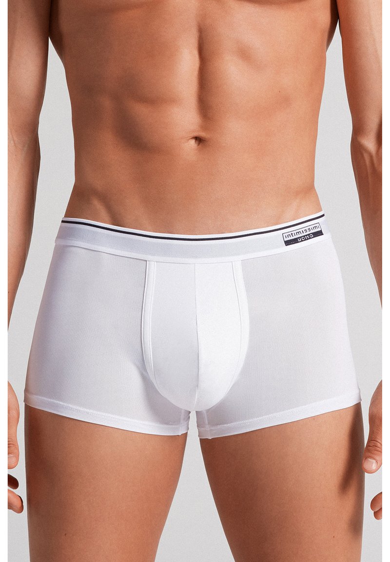 Mannelijk bovenlichaam met witte, aansluitende boxershorts met een merkband en een voorzak, staand tegen een neutrale achtergrond.