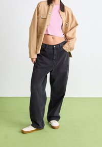 Beige spijkerjasje, zwarte wijde jeans en witte sneakers met bruine zolen; roze cropped top zichtbaar. Vlakke groene achtergrond.