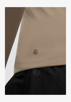 Primer plano de una camiseta de manga larga beige y pantalones negros, ambos con pequeños logotipos circulares cerca del dobladillo.