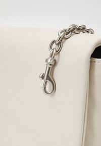 Detailní záběr stříbrného řetízku a spony připevněné k klopně krémové kožené kabelky.