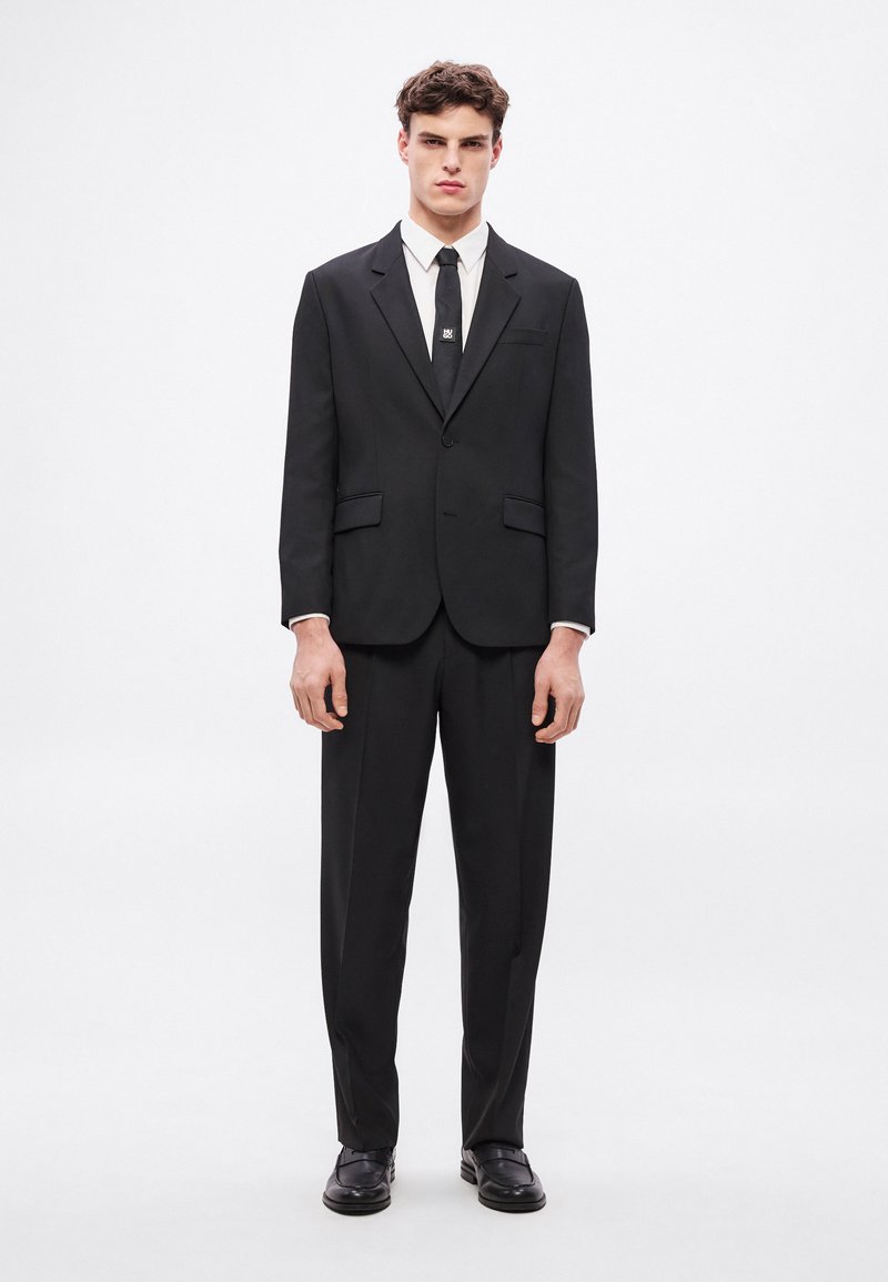 Czarny garnitur z marynarką na jeden guzik i szpiczastymi klapami, w komplecie z prostymi spodniami. Do kompletu biała koszula i czarny krawat.