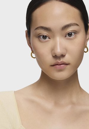 Pendientes de aro de oro con un diseño texturizado y ondulado. La modelo tiene la piel clara y lleva un top de canalé beige, con una piel suave y sin imperfecciones.
