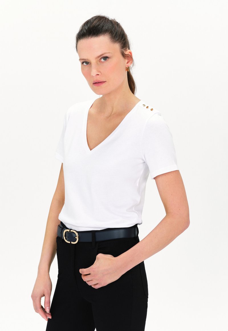 T-shirt bianco a V con maniche corte, con bottoni dorati sulla spalla sinistra, abbinato a pantaloni neri e una cintura nera.
