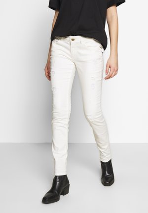 Osoba w białych, przetartych, obcisłych jeansach, czarnej bluzce i czarnych botkach na obcasie, stojąca na tle jednolitego białego tła.