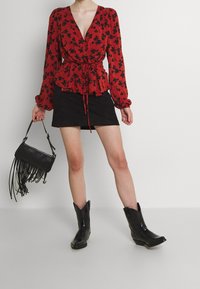 Blouse florale rouge à manches longues et à taille ceinturée, associée à une mini-jupe en suède noire et des bottes de cowboy noires. Sac à main frangé noir.