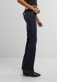 Jeans a gamba larga in denim scuro con vita alta, cuciture decorative sulle tasche e una leggera elasticità. Abbinati a stivali neri con tacco a punta.
