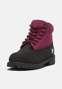 Botas de tobillo negras y burdeos, con materiales de ante y cuero, punta redonda, cuello acolchado y cordones, que presentan un logo circular en el lateral.