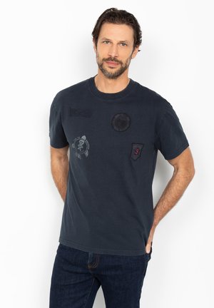 Homme aux cheveux courts et foncés avec une barbe, portant un t-shirt noir avec des patchs et un jean bleu foncé, debout les mains dans le dos.