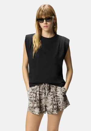 Femme aux longs cheveux blonds portant un haut noir sans manches, un short imprimé serpent, des boucles d’oreilles créoles et des lunettes de soleil noires en œil de chat, les mains dans les poches.