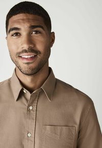 Camisa marr�ón de botones fabricada en tejido suave, con un collar, un bolsillo en el lado izquierdo del pecho y botones claros. El modelo posa con una expresión neutral.