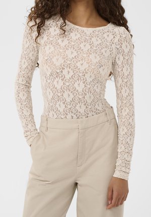 Vrouw met een crèmekleurige kanten top met lange mouwen, ingestopt in een beige broek met hoge taille, staat met één hand in de zak tegen een effen achtergrond.