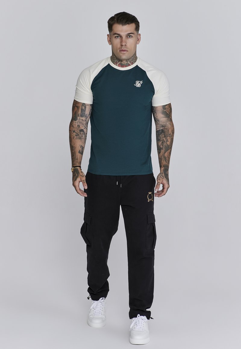 Teal short-sleeve shirt met crèmekleurige raglanmouwen en een logo op de borst, gecombineerd met zwarte cargo pants en witte sneakers.