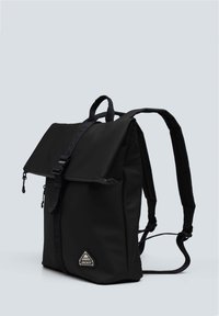 Mochila negra con cierre de solapa, fabricada en un tejido duradero. Cuenta con correas ajustables y un parche con un logo triangular. Textura suave.