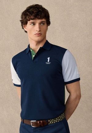 Jeune homme portant un polo bleu marine avec des manches blanches, un détail vert au col et une ceinture à motifs, debout devant un fond beige uni.