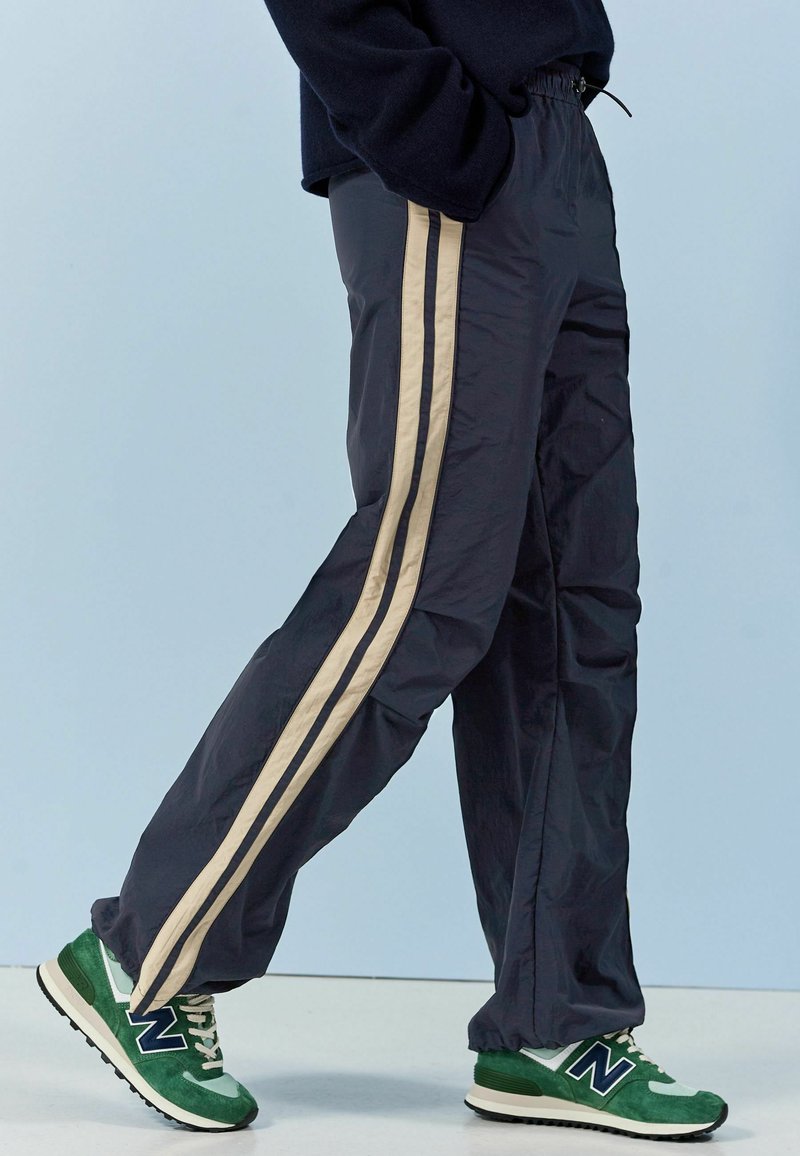 Navy jogginghousut, jotka on valmistettu kevyestä materiaalista ja joissa on beige sivuvaruste; yhdistetty vihreisiin ja valkoisiin New Balance -lenkkareihin.