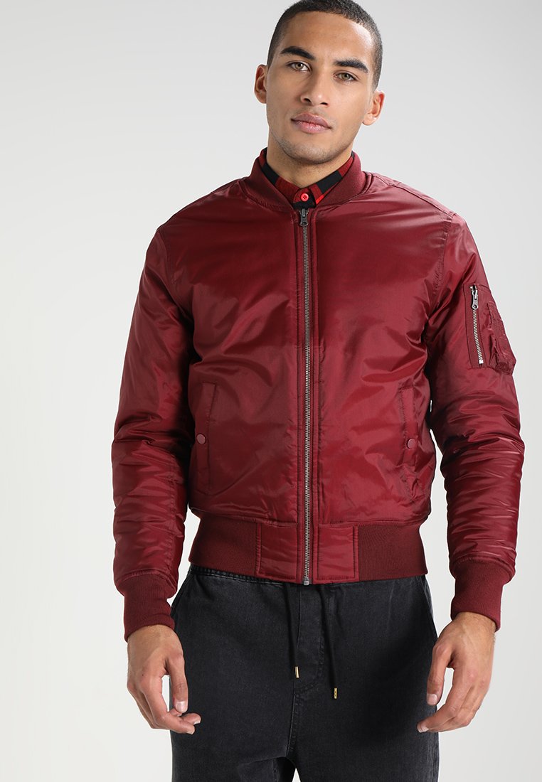 Giacca bomber bordeaux con chiusura a zip, polsini e orlo a coste. Presenta una tasca sul braccio sinistro. Materiale liscio e lucido.