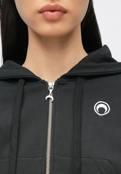 Casaco preto com fecho de correr prateado, apresentando um pequeno logótipo circular branco na área esquerda do peito. Feito de tecido texturizado.