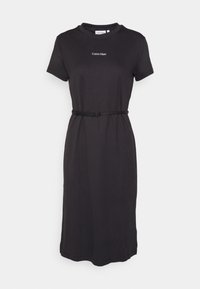 Robe midi noire à manches courtes avec logo Calvin Klein sur la poitrine et fine ceinture noire à la taille, présentée sur un fond uni.