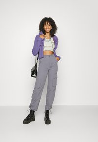 Lila, kurz geschnittene Strickjacke mit Kunstfellbesatz, graues gemustertes Crop-Top, graue Cargo-Hosen mit Taschen und schwarze Schnür-Plateau-Stiefeletten.