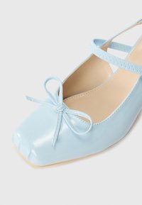 Ballerine in vernice blu chiaro con punta quadrata e fiocchetto decorativo sul davanti. Presenta una cinghia alla caviglia con finitura testurizzata.