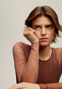 Top largo marrón de manga larga y escote redondo. La modelo descansa la barbilla en la mano, luciendo un anillo delicado. Textura suave y lisa, diseño minimalista.