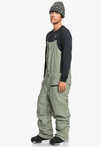 Salopette de ski en tissu vert clair avec des bretelles ajustables et des poches à fermeture éclair. Portée par-dessus un haut à manches longues noir et assortie avec des bottes noires.