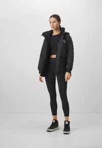Black quilted jacket with a hood, zipper closure, and side pockets, paired with black leggings and sneakers, all in a fitted design.