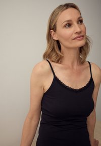 Donna con capelli biondi di media lunghezza che indossa un top nero con rifiniture in pizzo, guarda leggermente verso l'alto a destra contro uno sfondo semplice.