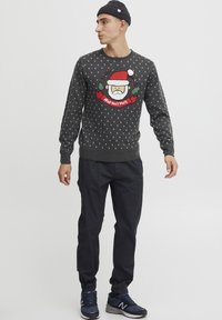 Pull de Noël en tricot gris avec un motif de Père Noël, des accents rouges et des pois blancs. Associé à un pantalon noir et des baskets bleu marine.