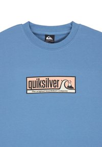 Camiseta de algodón azul claro con un logo rectangular en negro y rosa que incluye el texto "quiksilver" y un diseño de ola.