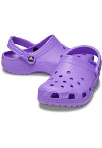 Zoccoli in plastica viola con apertura sul retro, fori di ventilazione e suole testurizzate. Presentano un logo circolare sul lato e una fascia rigata nella parte superiore.
