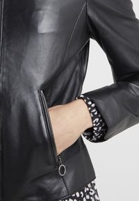 Černá kožená bunda s přiléhavým designem, vybavená kapsou na zip a vzorovanou podšívkou rukávu. Hladký povrch a minimální kovové doplňky.