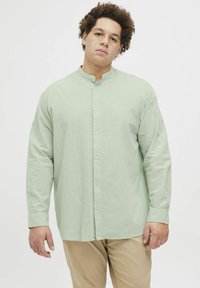 Camisa de manga larga verde claro con cuello mao, que presenta un cierre de botones, textura suave y corte relajado. Combinada con pantalones beige.