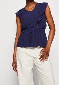 Námořnická modrá blůza s volány, ve výstřihu do V, s šňůrkami v pase a krátkými rukávy, spárovaná s krémovými širokými kalhotami. Hladká textilie.