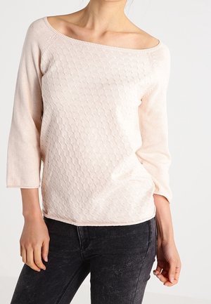 Kobieta ubrana w jasnoróżowy dzianinowy sweter o fakturze z rękawami do łokci i czarne spodnie na jednolitym białym tle.