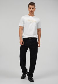 Camiseta blanca de algodón con el texto "Palm Spring" en dorado. Combinada con pantalones negros y zapatos negros. Modelo de pie frente a un fondo liso.
