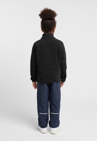 Veste en polaire noire avec col haut, associée à un pantalon cargo bleu marine comportant des bandes réfléchissantes, le tout complété par des chaussures blanches.