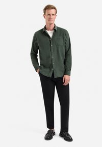 Donkergroene overhemd met knopen en een borstzak, gecombineerd met zwarte broek en zwarte instappers. Gladde stof met een relaxed fit.