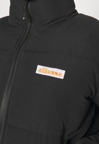 Veste matelassée noire avec fermeture éclair à l'avant et un patch rectangulaire blanc affichant le logo orange "ellesse" sur le côté gauche de la poitrine.