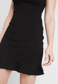 Svart figurnära klänning i elastisk tyg, med fransar i fållen och slät textur. Designen inkluderar en enkel, ärmlös siluett.