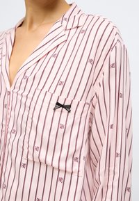 Cămașă roz deschis cu dungi verticale burgundy, având un buzunar la piept decorat cu o fundă neagră și logo-ul brandului într-un model repetat.